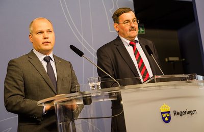 Morgans Johansson står till vänster bakom ett genomskinligt bord som det står "regeringen" på. Bredvid honom till höger står Lars-Erik Lövdén