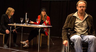 Foto från föreställningen. Hans Sjöberg sitter i förgrunden till höger och två andra personer sitter vid ett bord i bakgrunden. En har konduktörsmössa