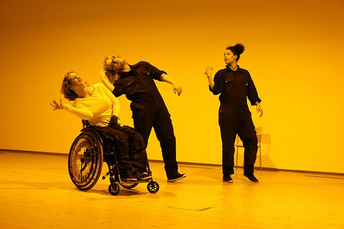 En bild från dansföreställningen Work. Tre personer dansar i ett gult rum. En använder rullstol två står.