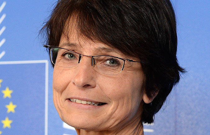 Porträtt av Marianne Thyssen mot en blå bakgrund där EUs flagga syns.