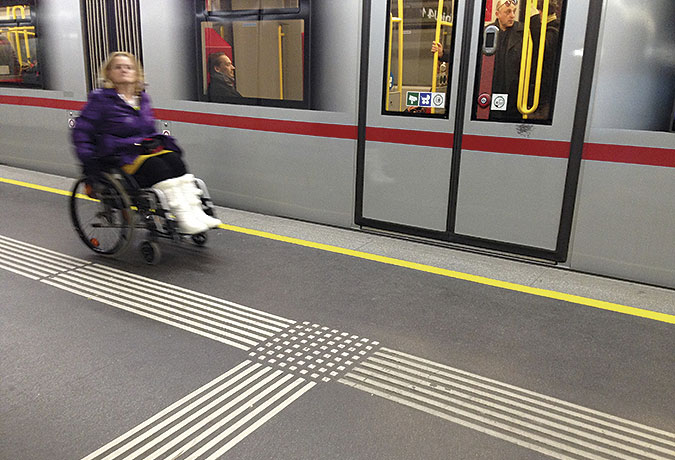 Kvinna i rullstol på perrong med ett grått tunnelbanetåg i bakgrunden och taktila ledstråk i golvet.