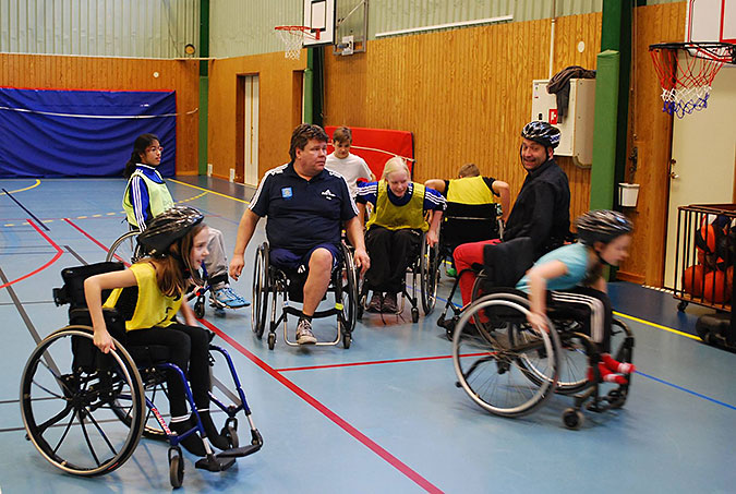 Barn kör rullstol i en idrottshall. Några har gula västar. Två vuxna är också med.