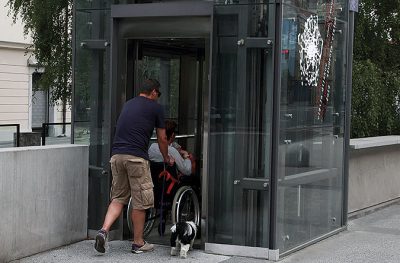 en person hjälper en person i rullstol att koma in i en inglasad hiss. En liten svartvit hund springer bredvid.