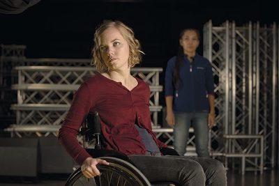 Ylva Andersson sitter i en rullstol i förgrunden. I bakgrunden står hennes assistent.