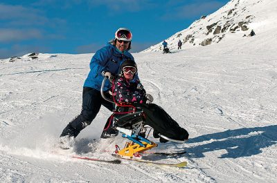 En skidlärare hjälper en sitski-åkare att ta sig ner för en skidbacke.