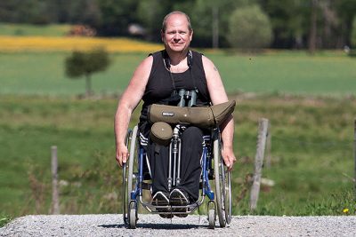 Pelle Aspegren sitter i en rullstol på en grusväg i ett öppet ängs- och åkerlandskap. I knät har han en fågelkikare med fodral och stativ.