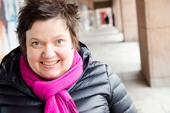 Sari Nykvist iklädd svart jacka och rosa halsduk. HOn sitter utomhus med en arkadgång i bakgrunden.