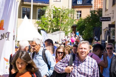 Många människor rör sig i solskenet på en gata i Visby som är kantad av rollups och tält från olika aktörer.