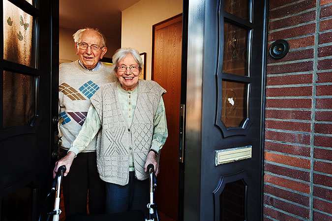 Ett äldre man och en en äldre kvinna står i dörren till ett hus och tittar mot oss. Kvinnan använder rollator.