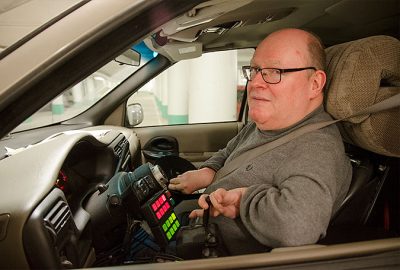 En man på förarplatsen i en bil. Här finns ingen vanlig ratt.Istället styr han med en spak och kontrollerar andra funktioner med särskilda knappar som syns i bilden.