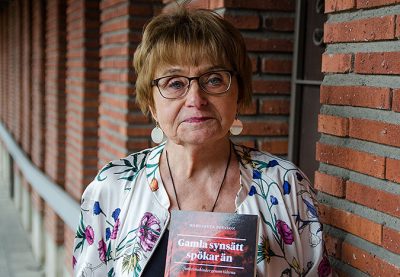Bild där Margarete Persson står vid en rödorange tegelvägg och håller i sin bok.