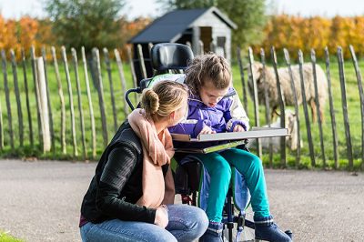 Ett barn i rullstol och en kvinna som sitter på huk bredvid. I abkrgunden står får bakom ett staket.
