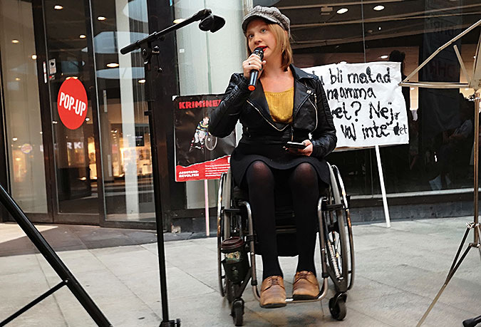 En kvinna sitter i rullstol och pratar i en mikrofon. I bakgrunden står några plakat lutade mot en glasvägg.