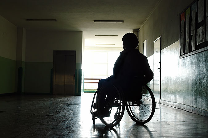 Person sitter i rullstol i en mörk och kal korridor där det enda ljuset kommer från ett fönster i bakgrunden.