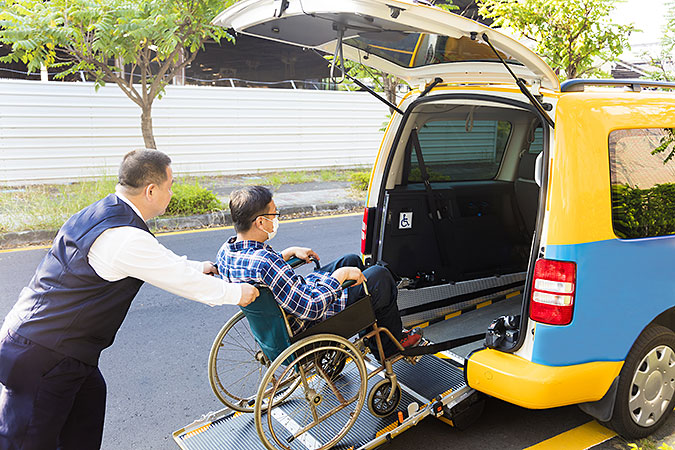 En färdtjänstchaufför hjälper en man i rullstol ombord i en liten gul och blå bil.