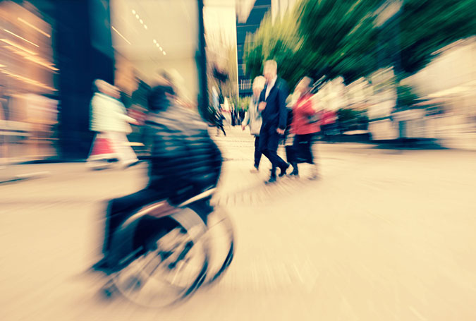 En person i rullstol tar sig fram på gatan bland andra människor. Bilden är tagen med en effekt som gör att det ser ganska stressigt ut.