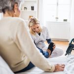 En yngre kvinna från hemtjänsten hjälper en äldre kvinna som sitter i en säng att ta på tofflorna.Bredvid sängen står en rullstol.