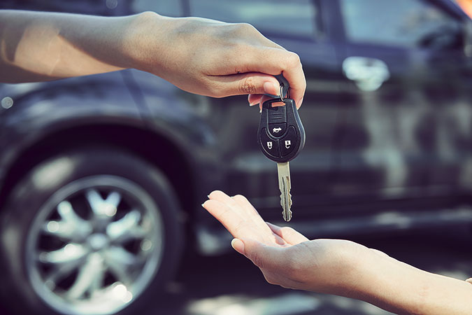 En person ger en bilnyckel till en annan person. I bakgrunden, som är oskarp, syns en mörk bil.