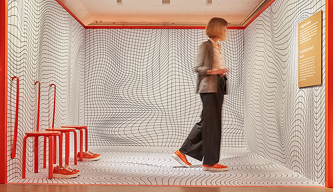 En person går i ett litet rum med ett oregelbundet rutnätsmönster på väggar och golv. Till vänster i bild står tre orangefärgade pallar med var sitt par orangefärgade skor.