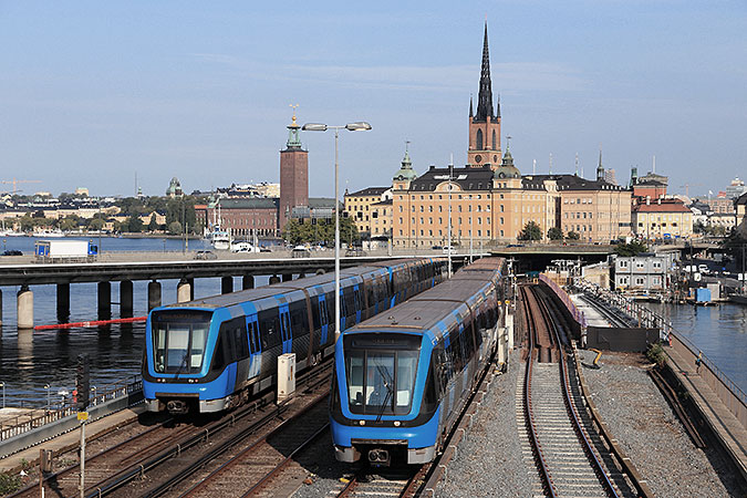 Två tunnelbanetåg kör på bron mellan slussen och gamla stan. I bakgrunden syns Stadshuset och riddarholmen.
