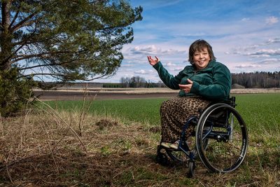En kvinna sitter i en rullstol vid kanten av ett grönt fält. I kanten av bilden finns ett träd.