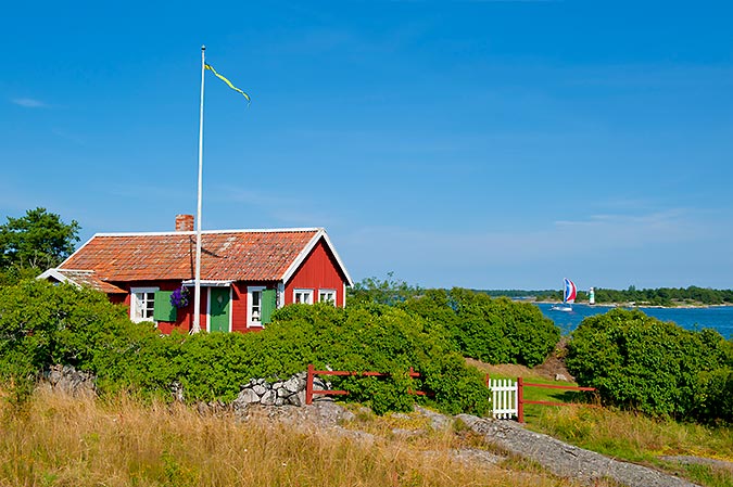 En röd stuga med vita knutar ligger inbäddad i grönska  alldeles bredvid kusten. I bakgrunden syns en segelbåt med spinnaker passera en lite fyr. Himmelen är blå.
