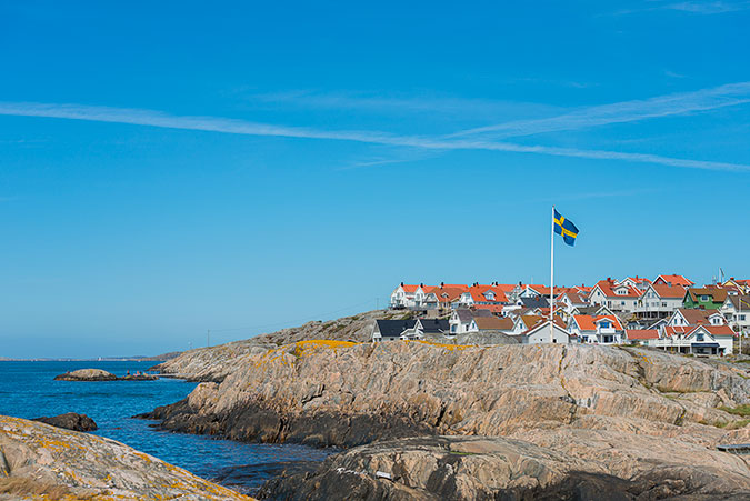 Bebyggelse på en kal klippö i skärgården. Blått hav, blå himmel och en flaggstång med svenska flaggan. 