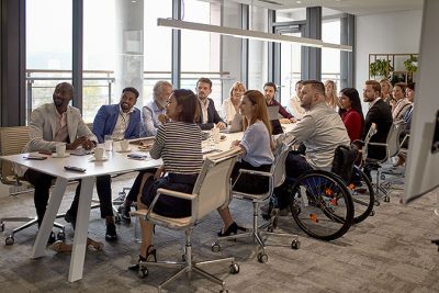 Ett dussin personer sitter i möte runt ett bord i ett konferensrum. De har sin uppmärksamhets riktad mot någon utanför bilden. En av personerna använder rullstol.