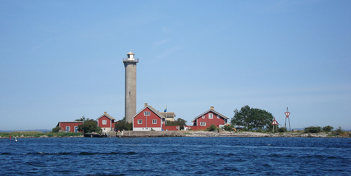En ö med en grå fyr och flera röda hus ligger med vatten runt om och en blå himmel övanför.