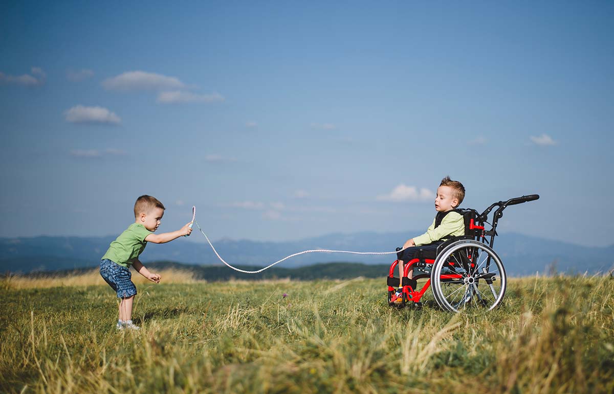 Två barn leker med ett hopprep på en vidsträckt äng. Ett av barnen sitter i rullstol, det andra står upp. Himlen är blå.