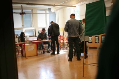 Tre personer köar i en vallokal för att avlägga sin röst. I förgrunden syns en röstningsskärm.