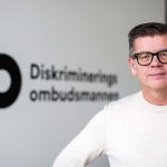 Bild på Lars Arrhenius som står framför en vit vägg där det står DO Diskrimineringsombudsmannen