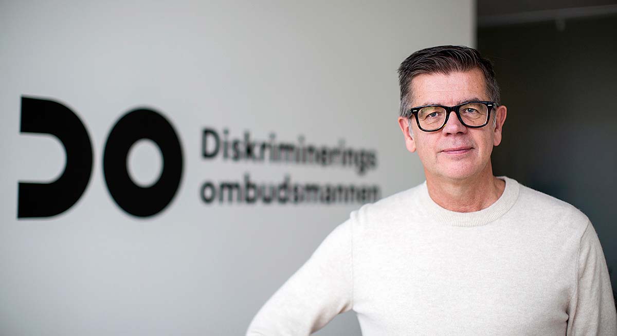 Bild på Lars Arrhenius som står framför en vit vägg där det står DO Diskrimineringsombudsmannen