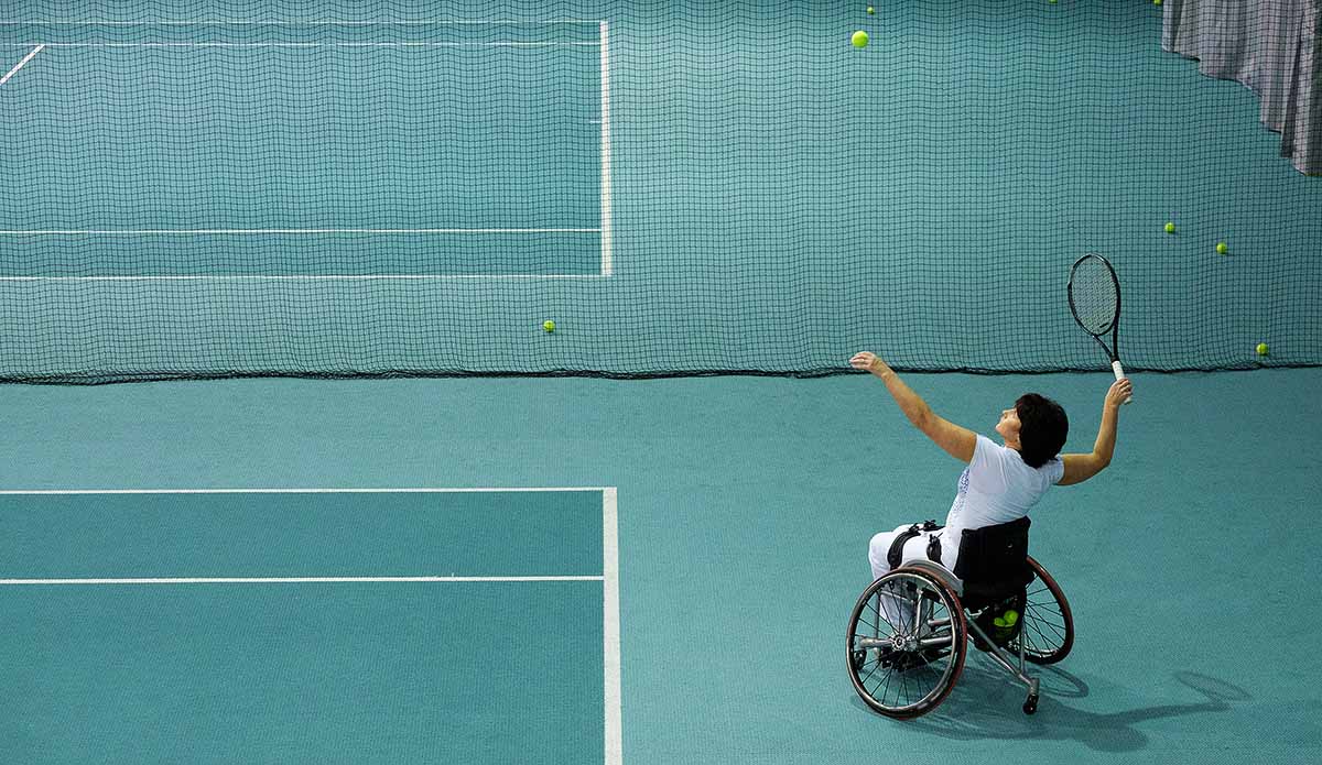 En kvinna i rullstol slår en serv bakom baslinjen på en tennisplan där golvet går i olika turkosa färger.
