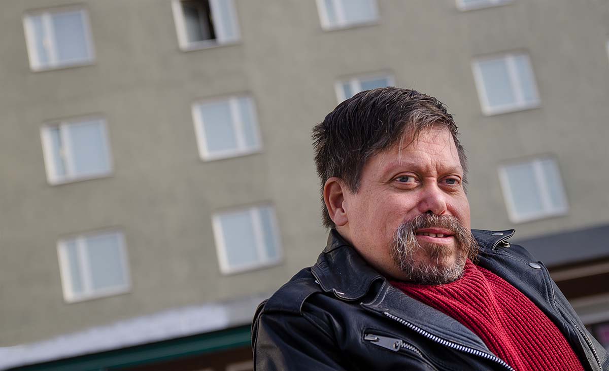 Bild av Patrik Forslin där man ser honom framför ett grått flervåningshus med många fönster.