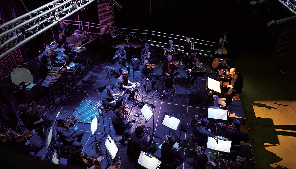 En orkester med stråkar och slagverk på en mörk scen. Till höger står dirigenten. I mitten sitter två personer som spelar på var sin iPad.
