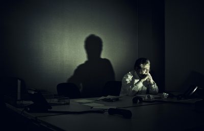 Deppig bild på man som sitter vid ett skrivbord i mörkret. Ljuset från den enda lampa kastar hans skugga på väggen bakom.