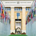 Rader med flaggstänger, med flaggor från olika nationer, står framför en ståtlig byggnad som den står United Nations – Nations Unies på.
