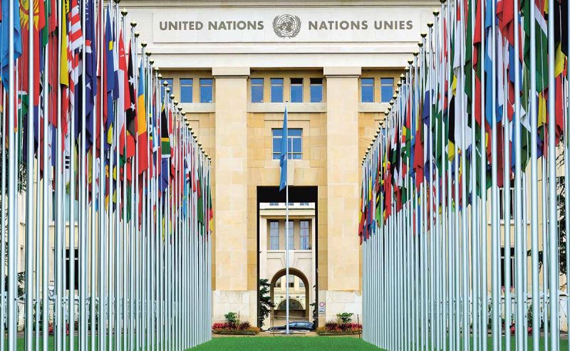 Rader med flaggstänger, med flaggor från olika nationer, står framför en ståtlig byggnad som den står United Nations – Nations Unies på.