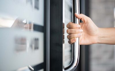 En hand greppar om ett dörrhandtag i rostfritt stål som sitter på en glasdörr.