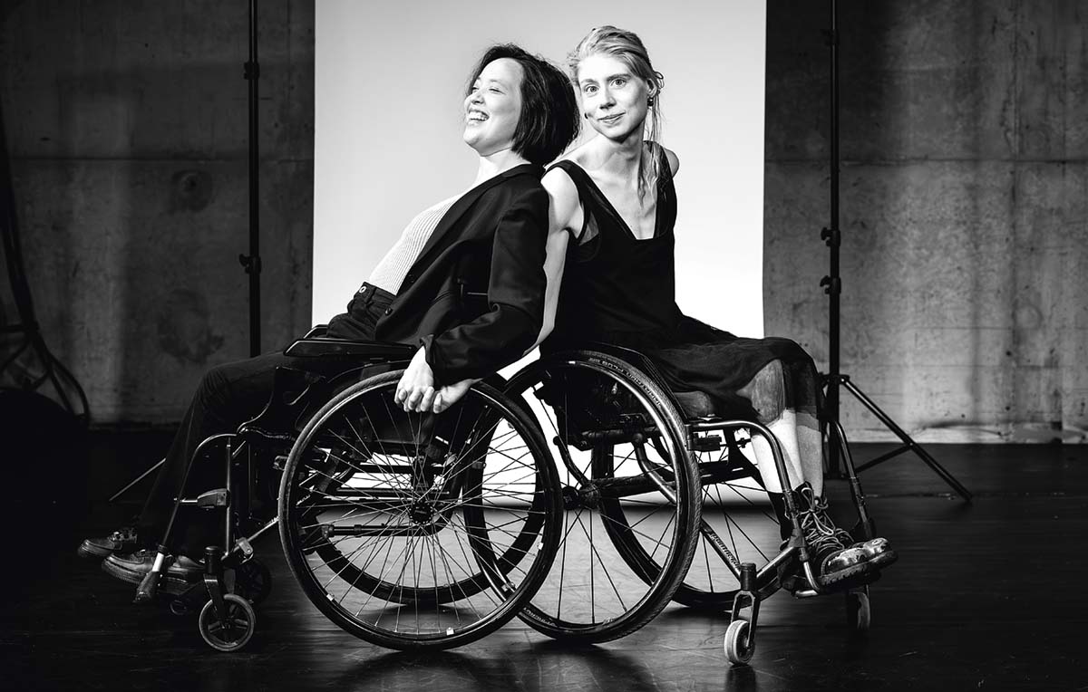 Två kvinnor i var sin rullstol sitter med ryggarna mot varandra. Den ena tittar in i kameran, den andra ser ut att skratta med slutna ögon.