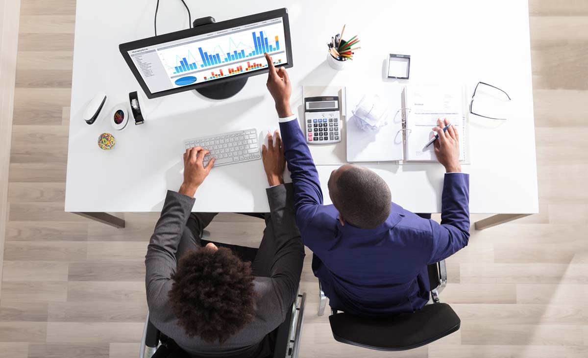 Bild tagen uppifrån på två personer som sitter vid ett vitt bard, med dator, papper och en miniräknare. En av personers använder rullstol