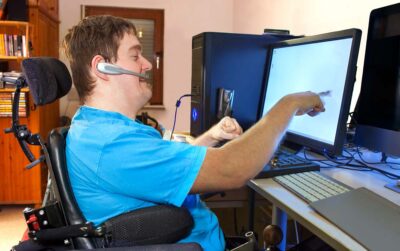 En man i rullstol sitter vid datorn med ett headset på. En bokhylla i bakgrunden.