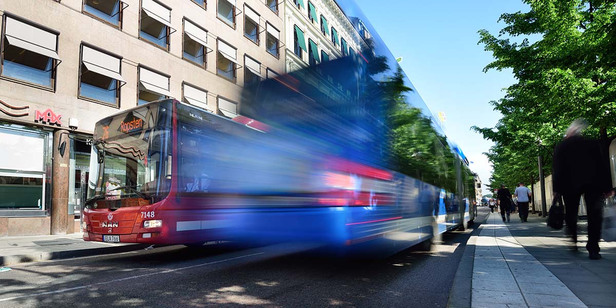 Två stadsbussar, en röd och en blå, möts på en smala gata med hållplatser i stan.